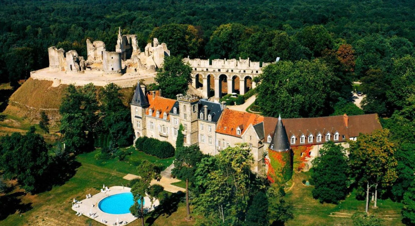 An aerial view shows a large, historic castle with red-tiled roofs and a swimming pool, surrounded by lush green trees and a forest.