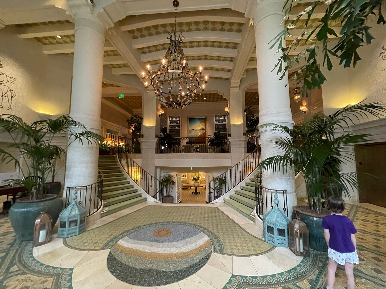 A grand, luxurious lobby features a central chandelier, sweeping green staircase, large columns, lush plants, and a mosaic floor.