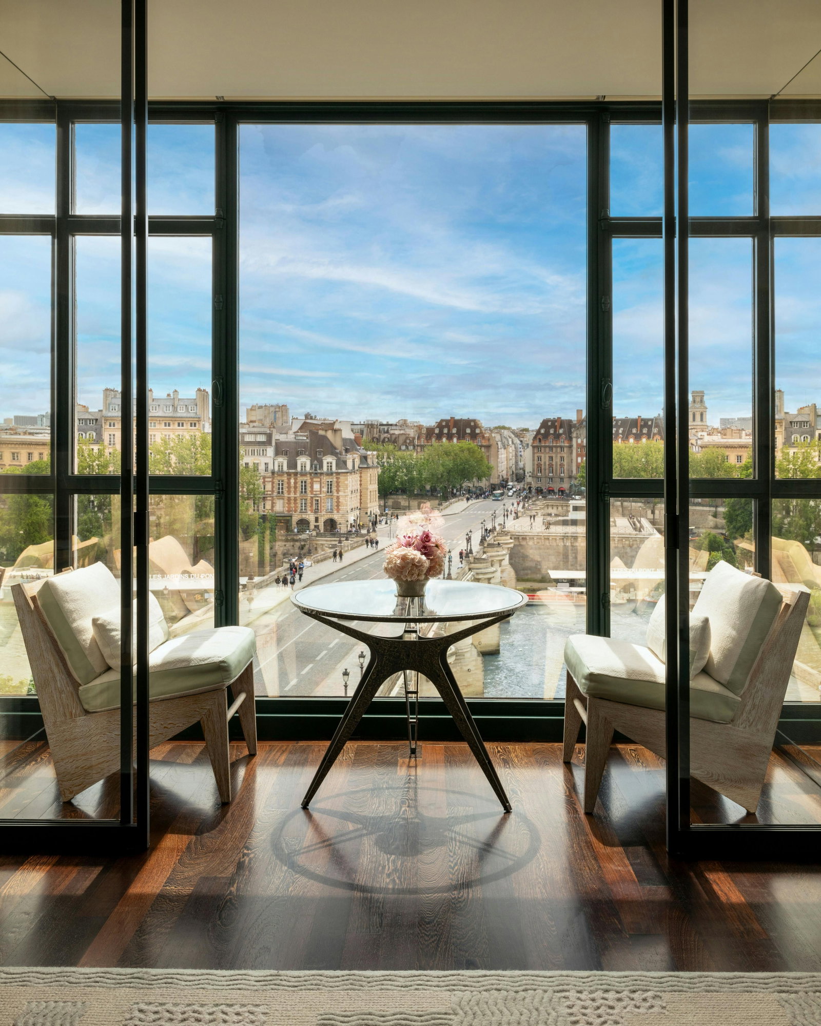 A room with large windows overlooking a city, river, and bridge, featuring two armchairs and a glass-top table with flowers.