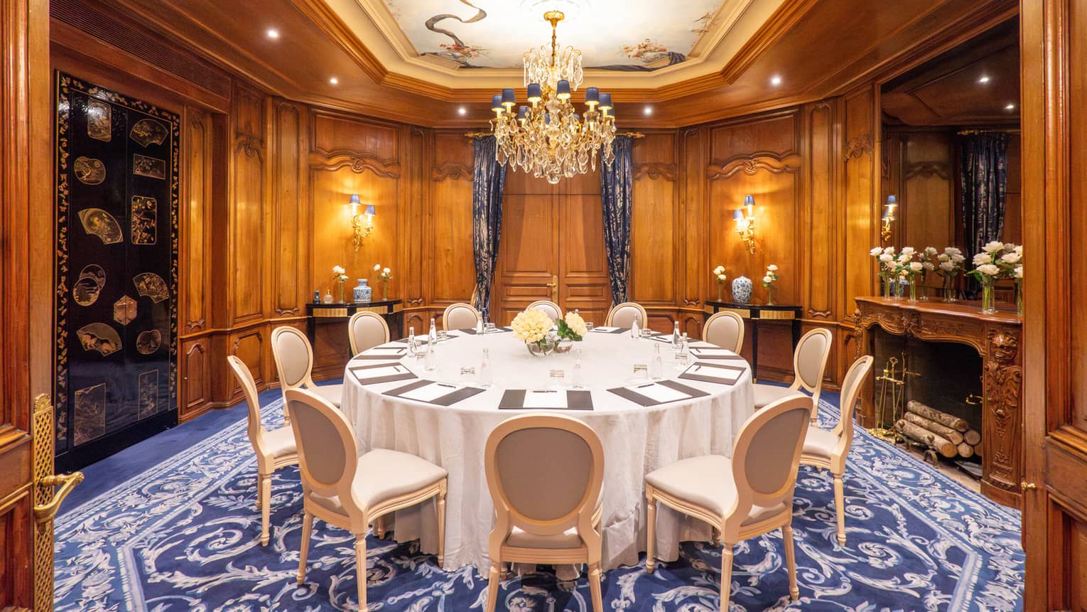 A luxurious meeting room with wood-paneled walls, a large round table with chairs, and an ornate chandelier.