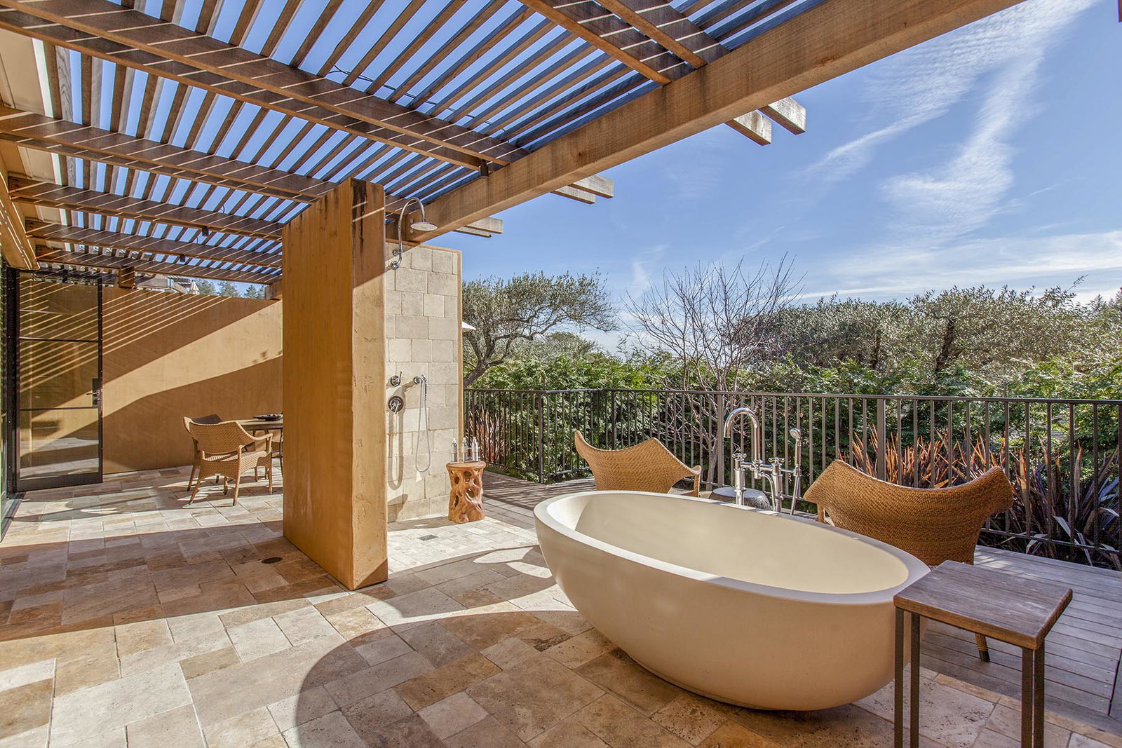 An outdoor patio with a wooden pergola overhead, featuring a freestanding bathtub, an outdoor shower, and a dining area with wicker chairs, all set.