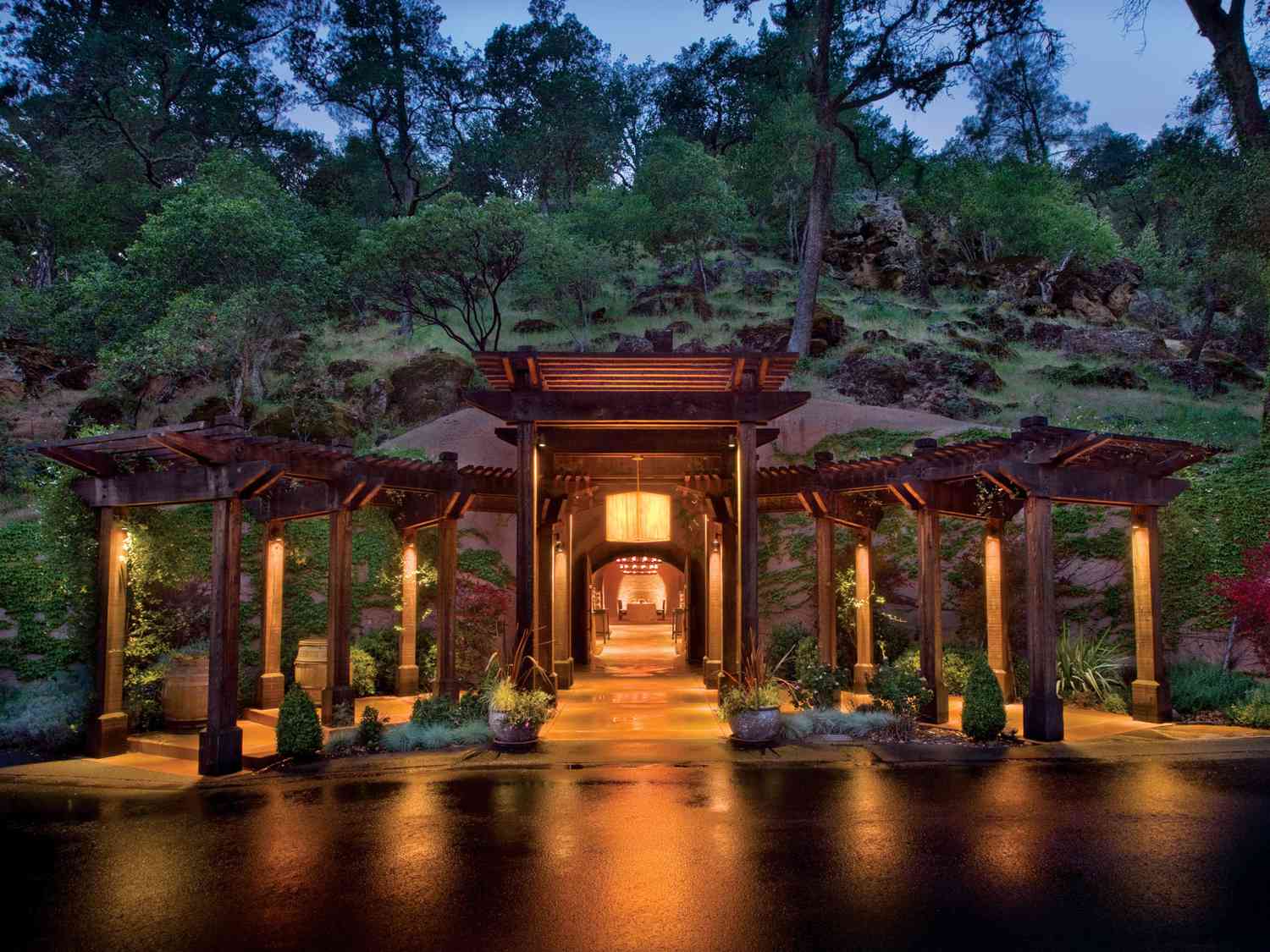 An illuminated wooden pergola entrance leads to a luxury hotel nestled into a lush, tree-covered hillside at dusk.