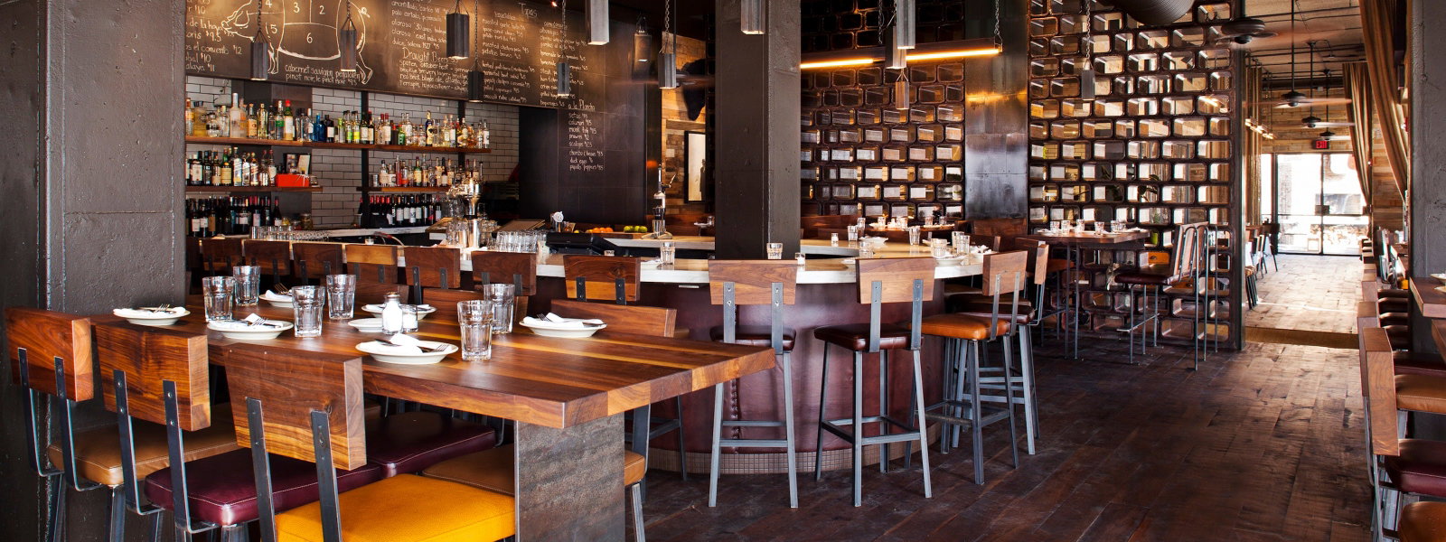 Interior of Barcelona Wine Bar in Inman Park, Atlanta, showing walnut communal tables set with white plates, a curved marble bar with metal stools, a perforated terracotta block wall, and chalkboard menus listing tapas and Spanish wines.