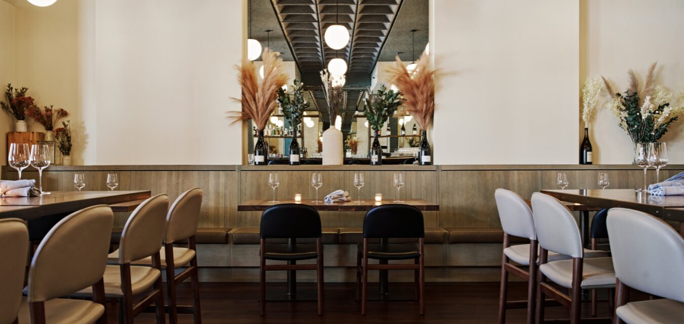 Empty dining room at Madeira Park Atlanta with oval tables, rounded upholstered chairs, a wood-paneled banquette, large antiqued mirror, and dried pampas grass decor.