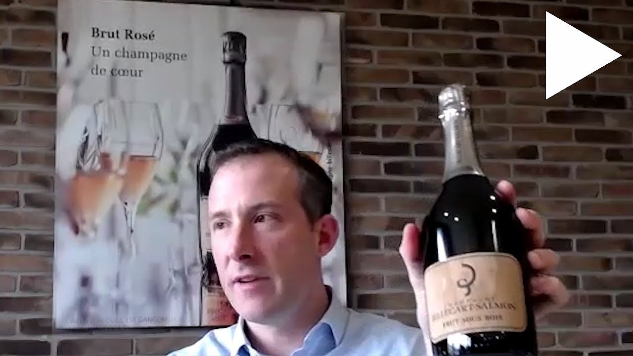 A man in business attire holding a bottle of Billecart-Salmon champagne in front of a brick wall with a Brut Rosé poster; the source page explicitly n