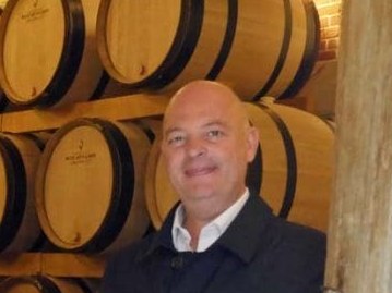A bald man wearing a dark blazer and white shirt smiles at the camera while standing in front of wooden wine or spirits barrels in what appears to be a cellar or aging room.