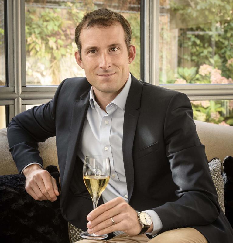 A man wearing a dark blazer and white shirt sits holding a champagne flute, with large windows showing greenery in the background.
