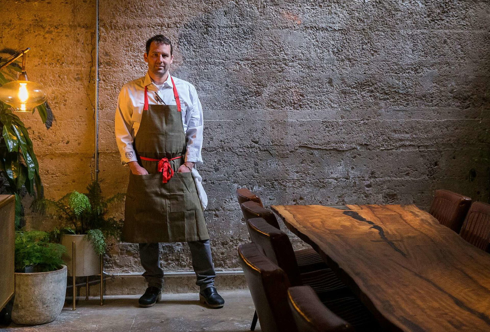 A man wearing a light blue shirt and gray apron stands in a restaurant interior with rustic stone walls, potted plants, and wooden dining furniture.