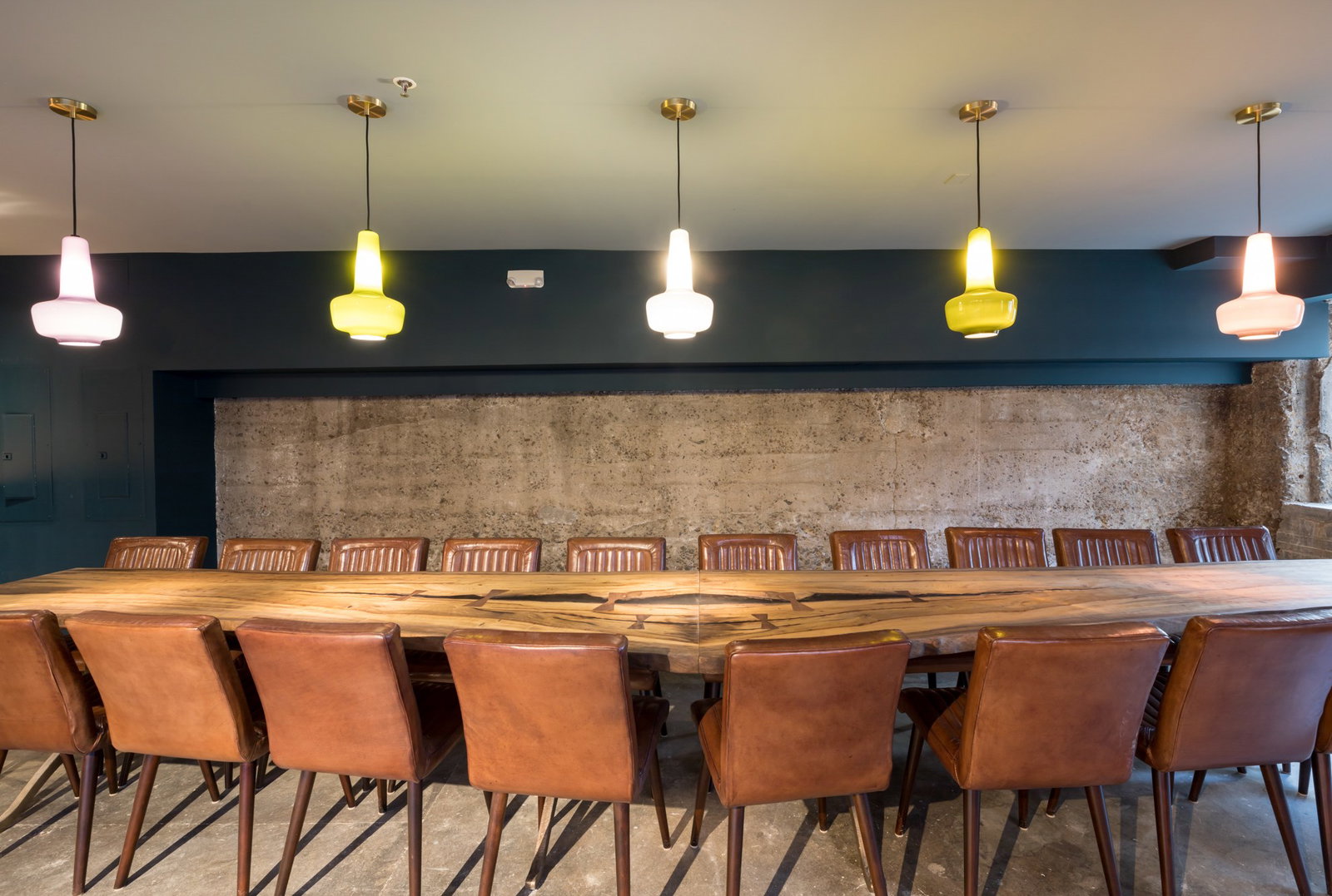 A modern private dining room with a long wood table, brown leather chairs, five hanging pendant lights in varying shades, and a textured brick wall backdrop.