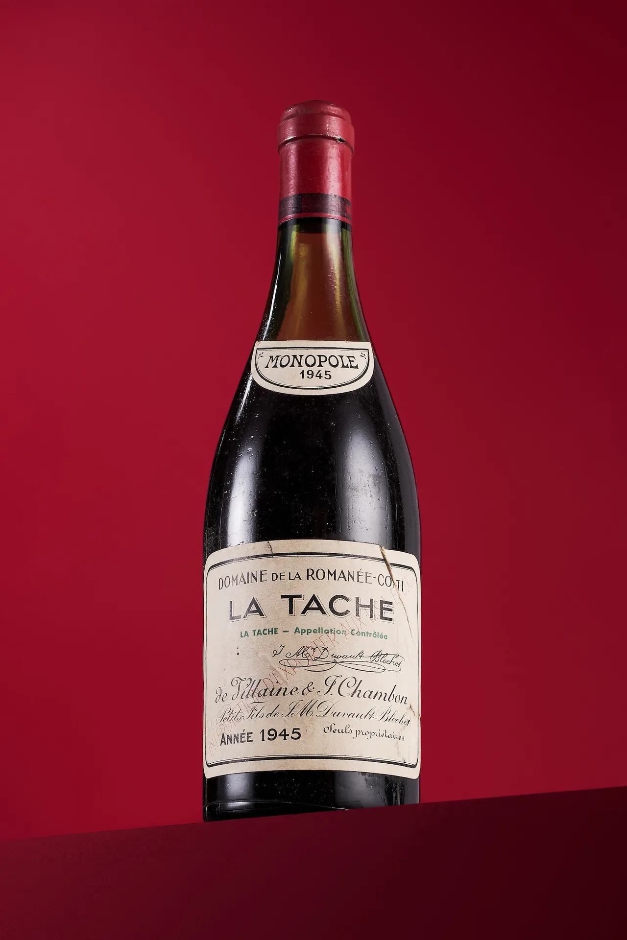 A bottle of Domaine de la Romanée-Conti La Tâche 1945 photographed against a red background; the label clearly shows 'LA TACHE', 'ANNÉE 1945', and 'MO