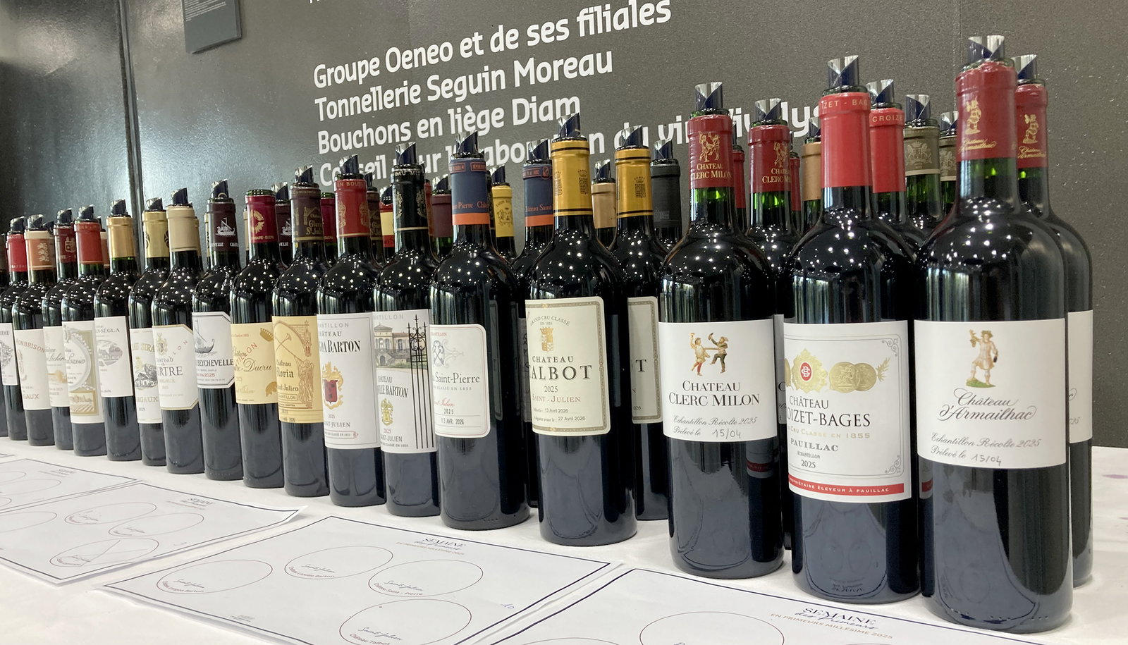 A row of red wine bottles, each with a label, stands on a white table with tasting sheets. The labels show names like "Château Talbot," "Château"