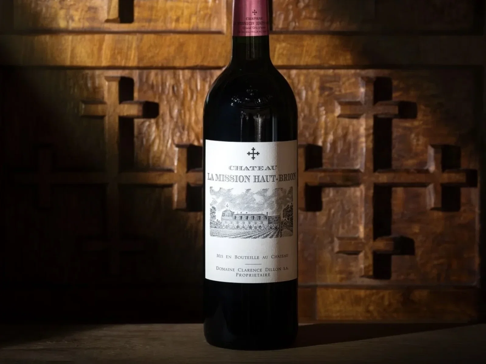 A bottle of Chateau La Mission Haut-Brion red wine stands upright against a dark wooden background with carved cross patterns.