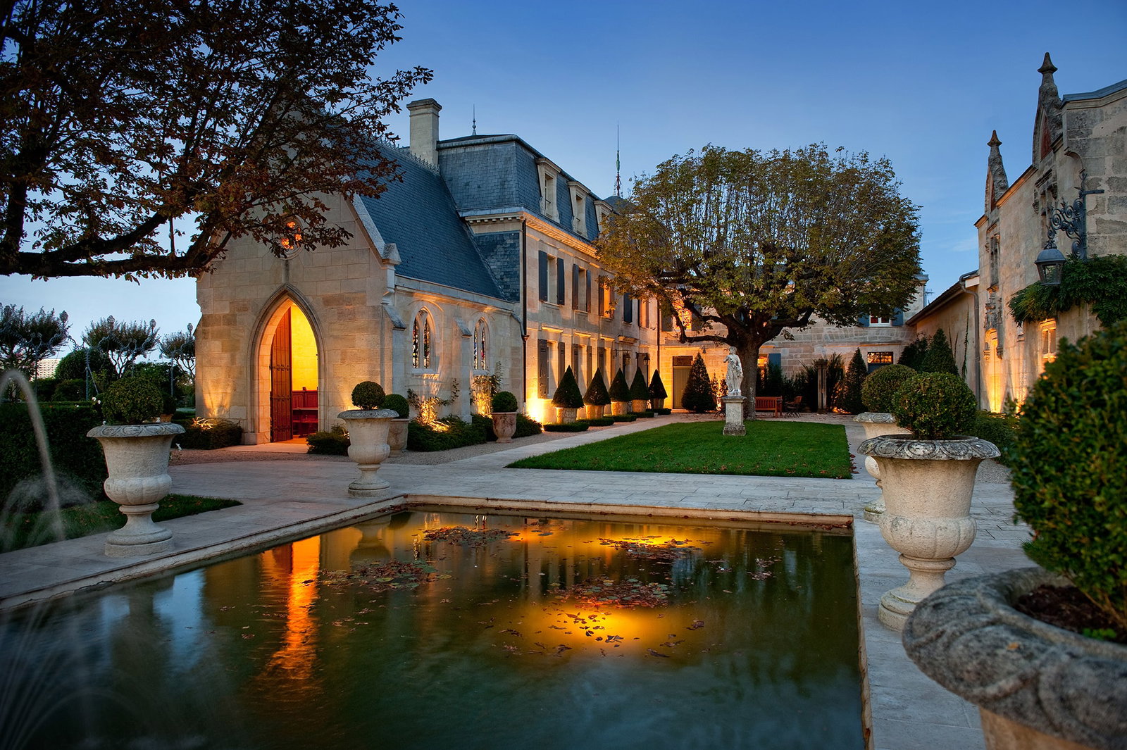 The illuminated stone facade of Chateau La Mission Haut-Brion at dusk, with a reflecting pool and manicured gardens in the foreground.