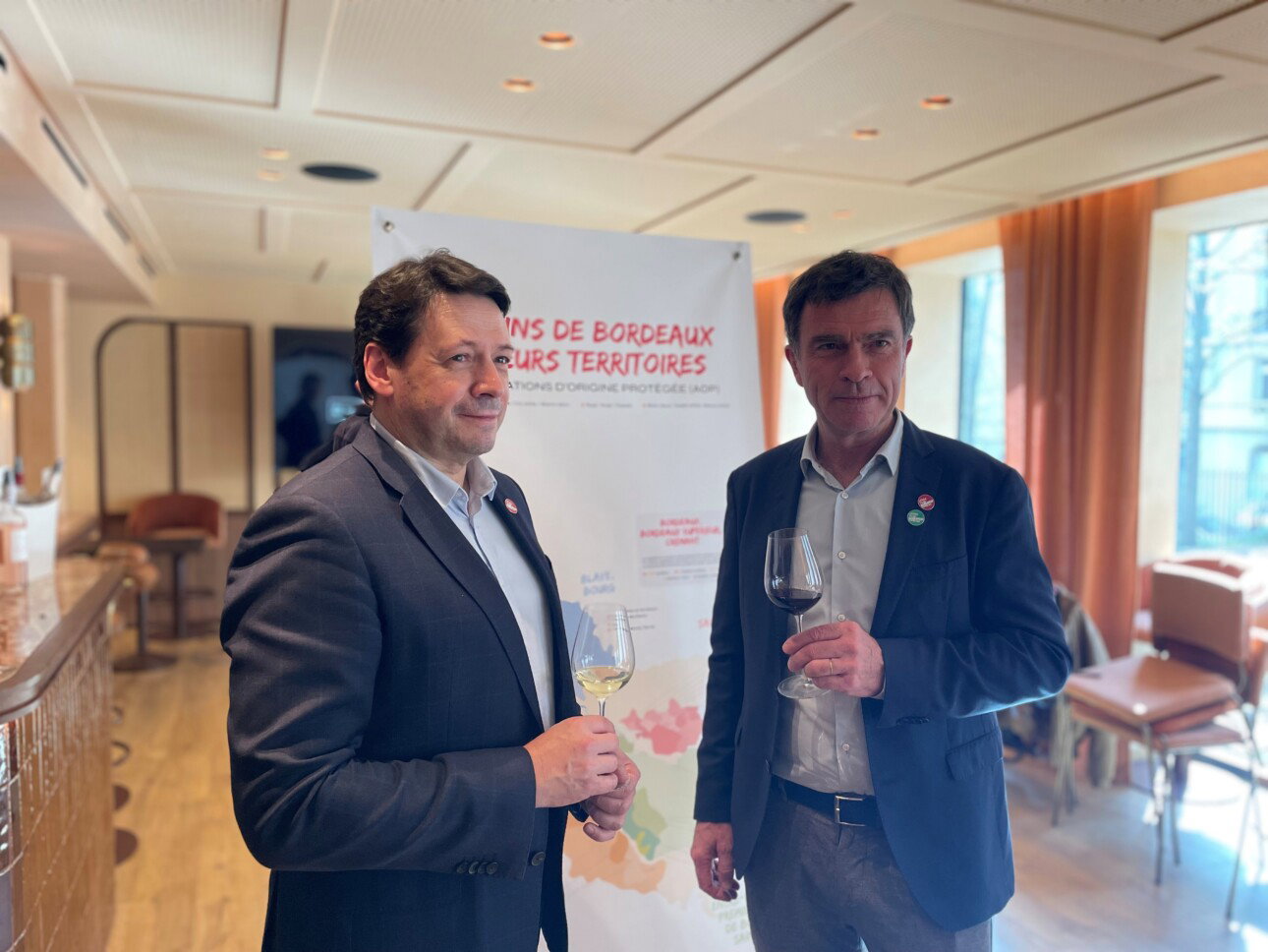 Two men in blazers hold wine glasses at a Vins de Bordeaux AOP promotional event, standing before a banner reading Vins de Bordeaux Leurs Territoires.