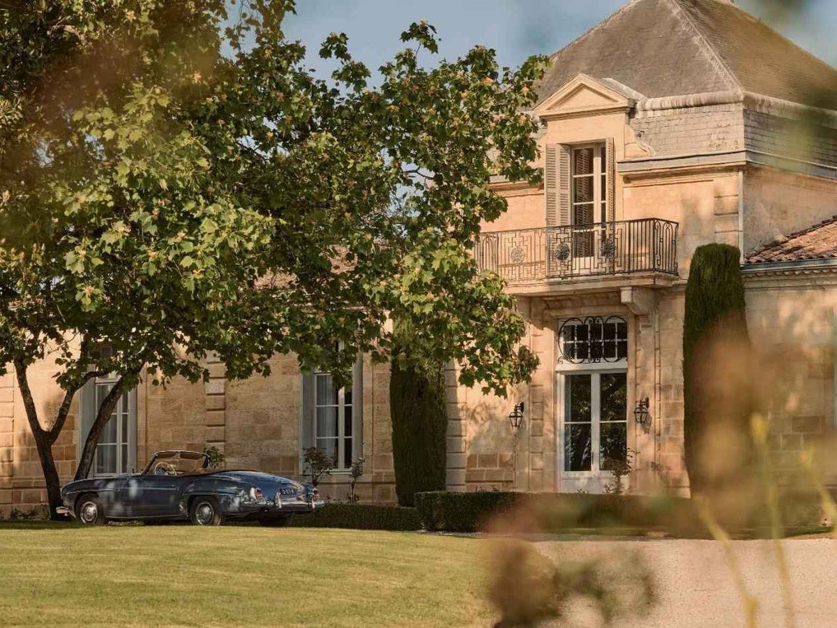 A restored 19th-century French château with a classic blue vintage car parked on the gravel drive, surrounded by trees.