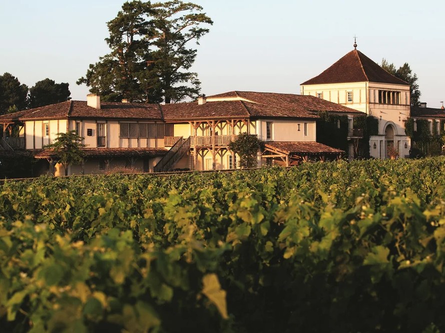 An exterior view of a low-rise estate building with terracotta-tiled roofs, a wooden balcony, and an octagonal tower amid vineyards.