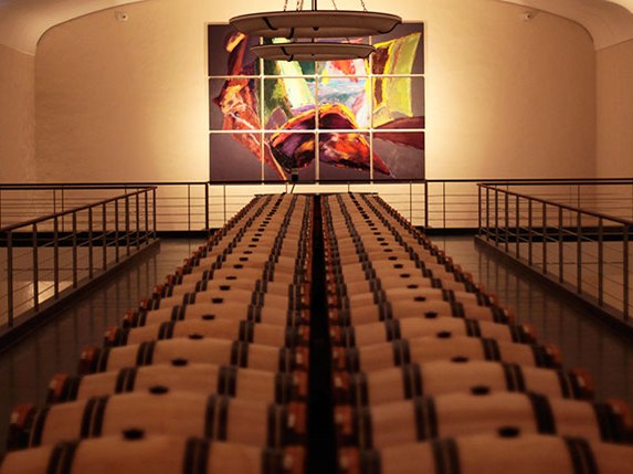Rows of oak wine barrels recede into a dimly lit cellar, with a large, colorful abstract painting on the far wall.
