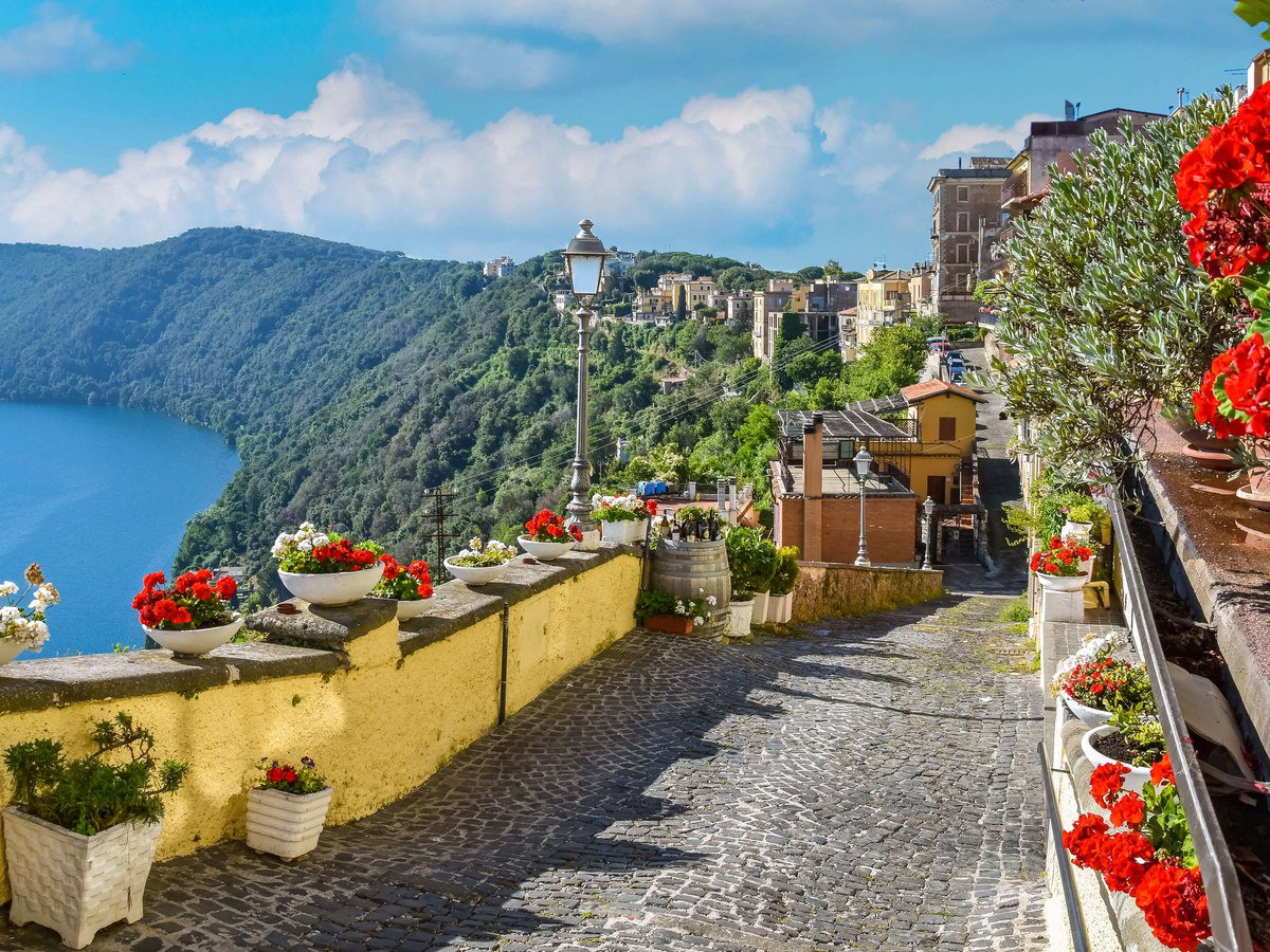 A cobblestone street in Castel Gandolfo, lined with potted red and white geraniums, overlooks the deep blue Lake Albano and lush green hills.