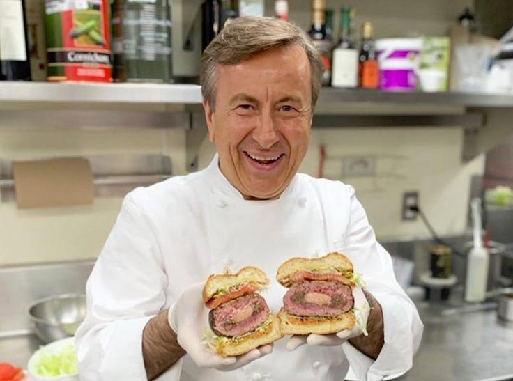 Chef Daniel Boulud in white chef's coat holding two DB Burgers with visible foie gras-stuffed patties in a professional kitchen; source page explicitl