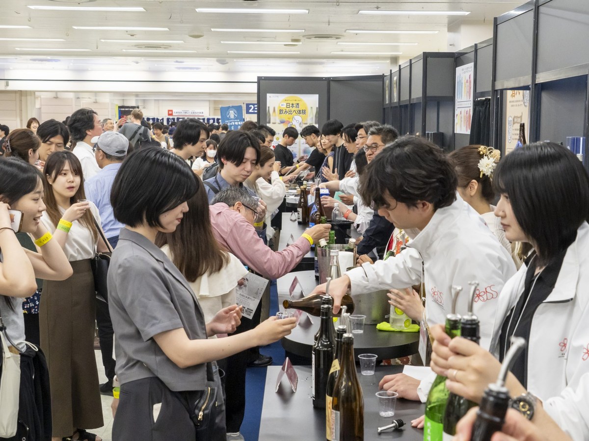 A crowded sake tasting event in Japan with many attendees sampling beverages at multiple booths, with visible Japanese signage.