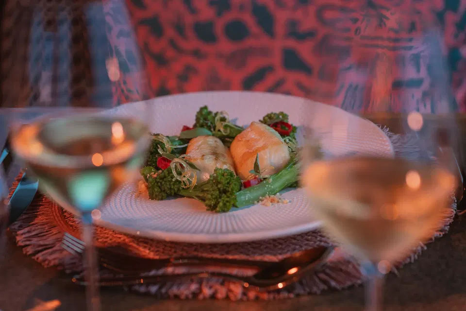 A close-up shot of a white plate with two pieces of fish, broccoli, and red peppers, with two wine glasses in the foreground.
