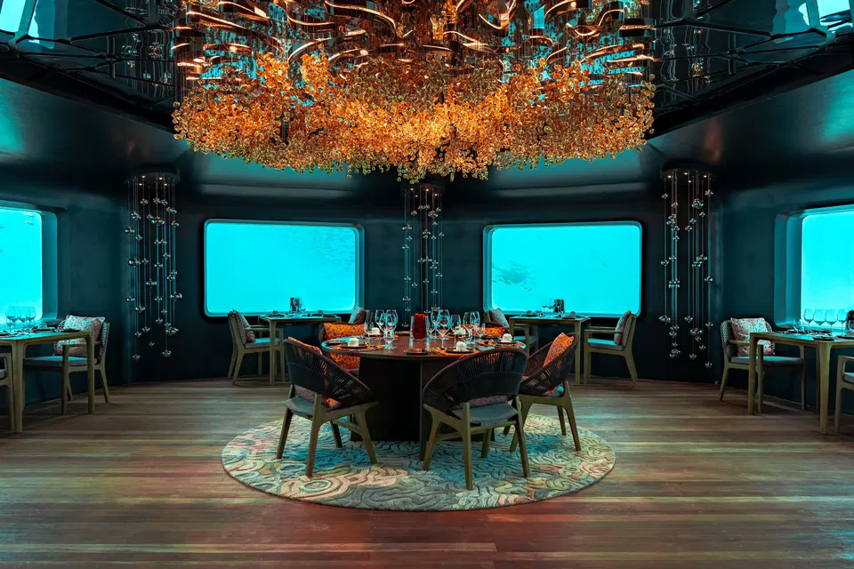 An underwater restaurant with a large, ornate chandelier made of golden, leaf-like crystals hanging from the ceiling. The room has dark walls and