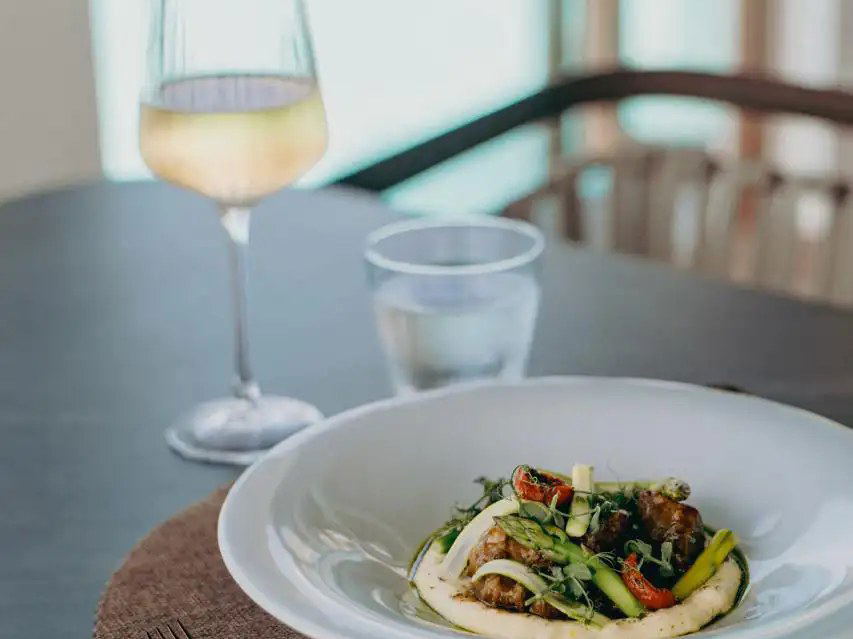 An elegantly plated dish with meat, asparagus, roasted tomatoes, and microgreens on a white plate, next to a glass of white wine.