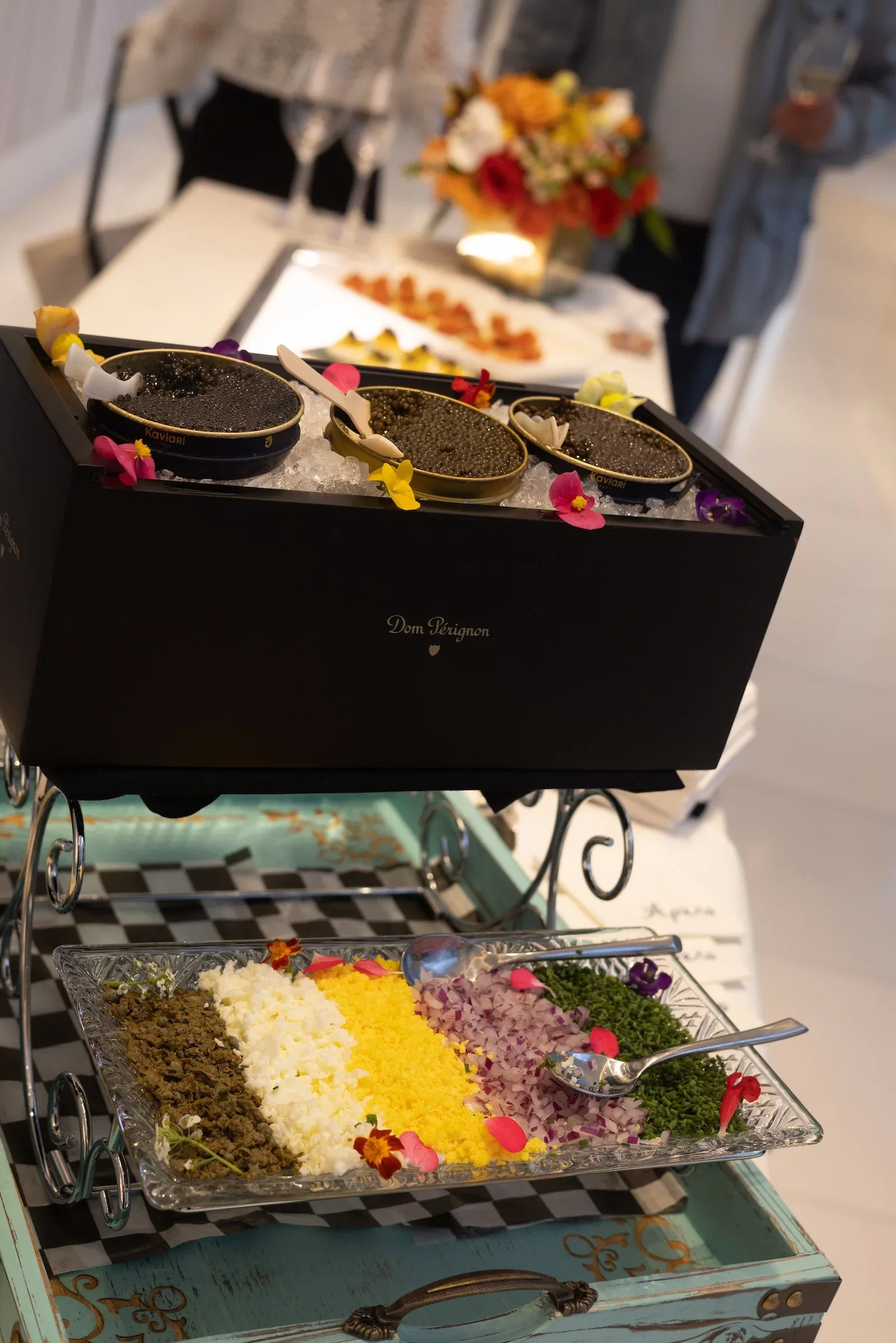 Elegant caviar service presentation with Dom Pérignon branded box containing three tins on ice, accompanied by colorful garnish tray and floral arrang