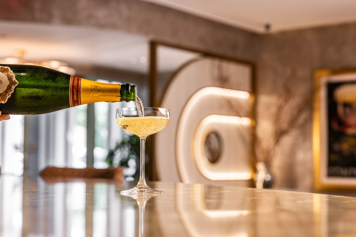 A bottle of champagne pours golden sparkling wine into a coupe glass on a polished counter in a sophisticated interior space.