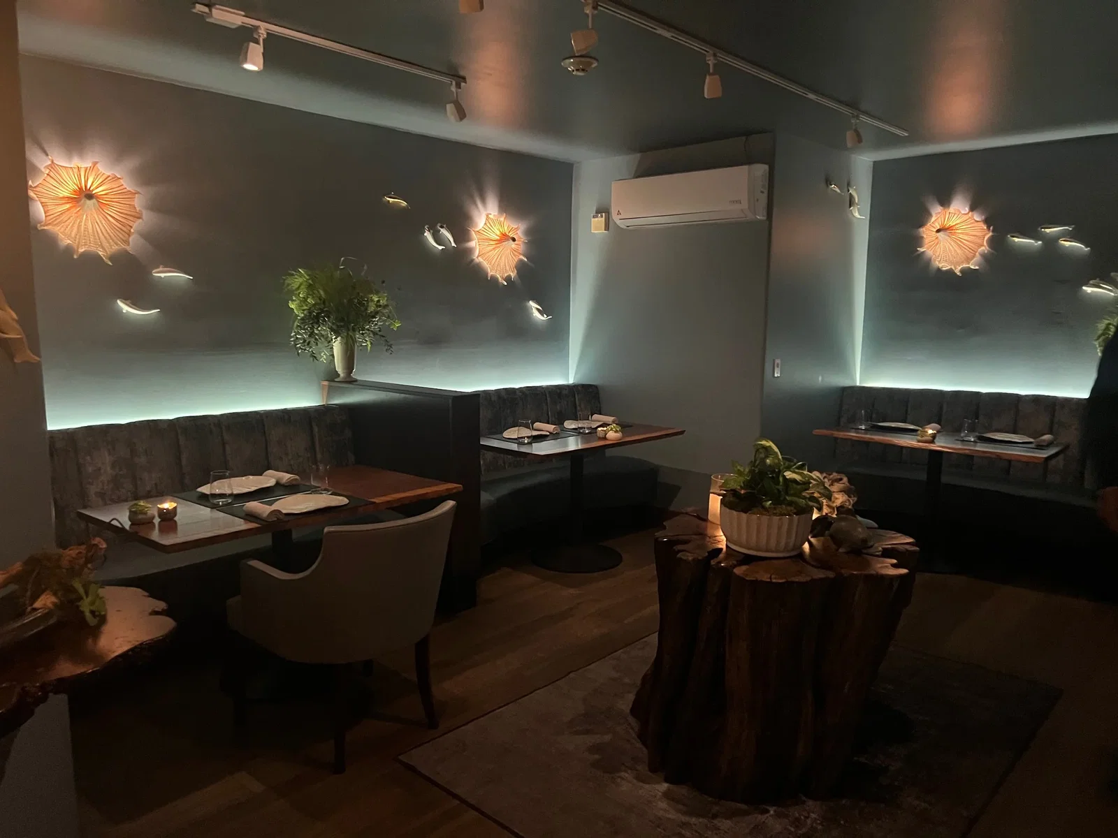 An upscale restaurant dining room with dark velvet booth seating, wooden tables set for service, decorative wall lighting fixtures resembling sea urch