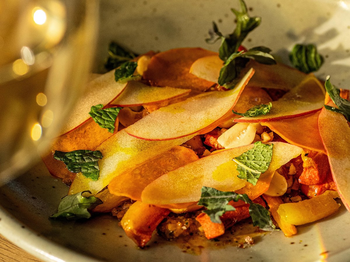 A beautifully plated tasting-menu course in a ceramic bowl featuring thin-sliced fruit (likely apple or peach), fresh mint leaves, toasted nuts, and d