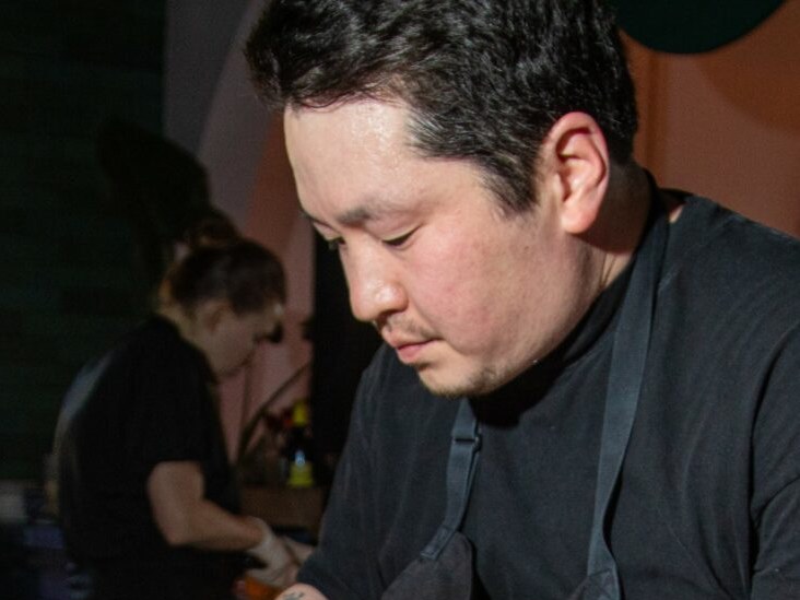 A man in a dark shirt focuses downward while wine tasting, with another person visible in the background at what appears to be an indoor venue.
