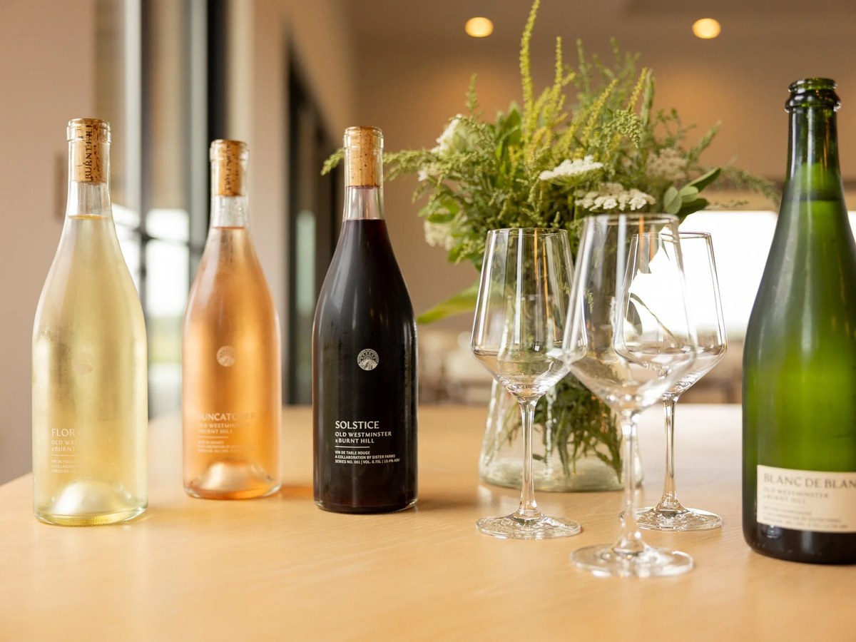 A styled counter display of four wine bottles (including a green-glass 'Blanc de Blancs' and 'Solstice Old Westminster x Burnt Hill' labeled bottles)