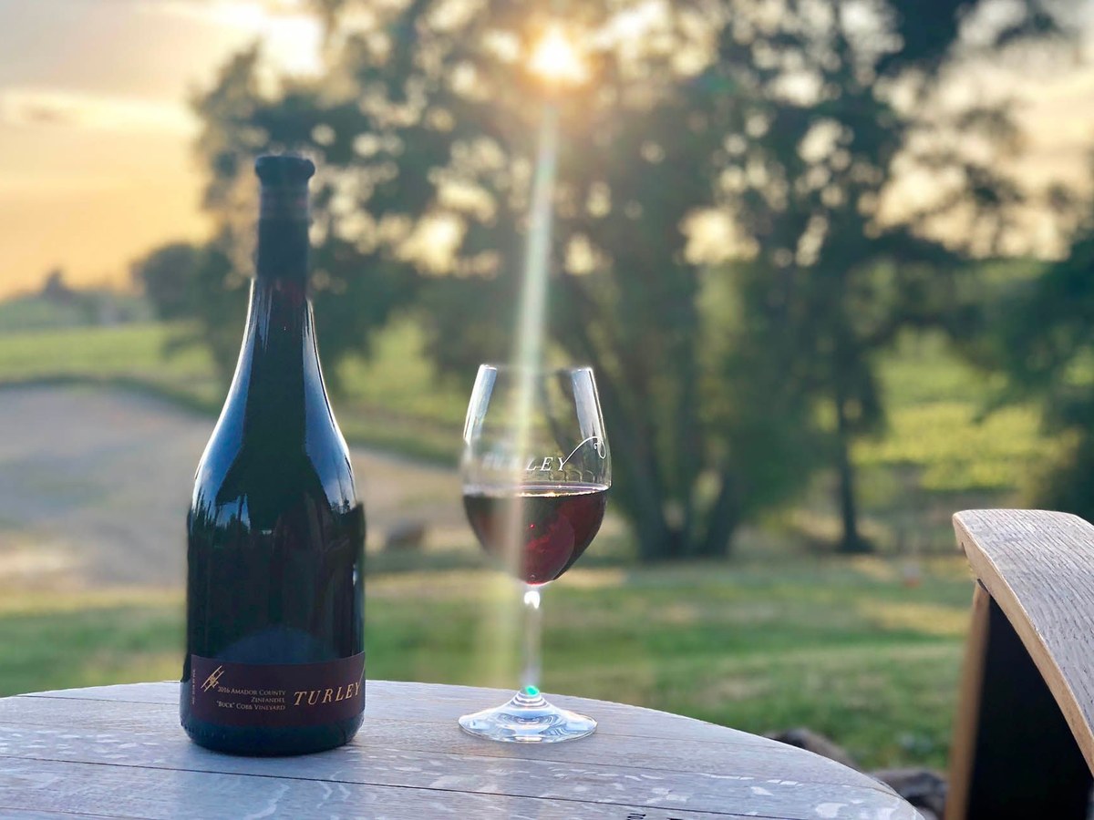 A bottle of Turley Zinfandel wine and a glass of red wine sit on an outdoor table with a vineyard and trees in the background at sunset.