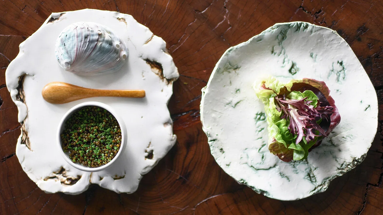 Abalone shell, wooden spoon, and a bowl of green caviar on a white plate, next to a plate with lettuce wraps.