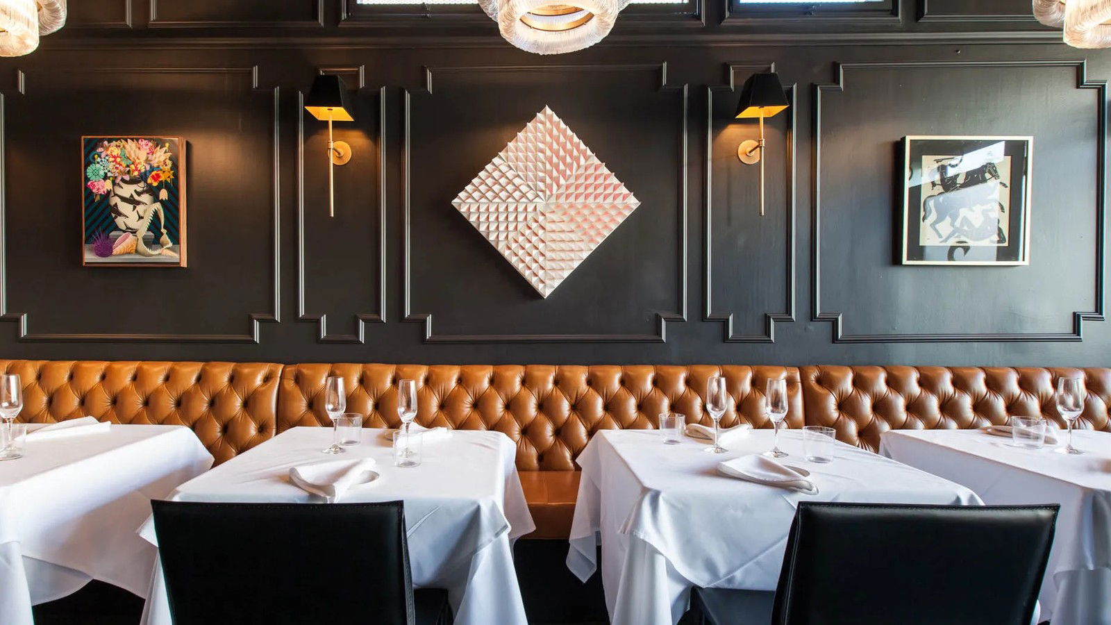An upscale restaurant dining room with dark walls, decorative molding, contemporary artwork, wall sconces, and tufted tan leather banquette seating.