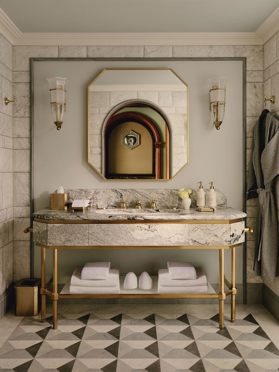 A luxury hotel bathroom vanity with marble countertop, brass fixtures, geometric floor tiles, and Art Deco-inspired design; the source page (Hotel Des