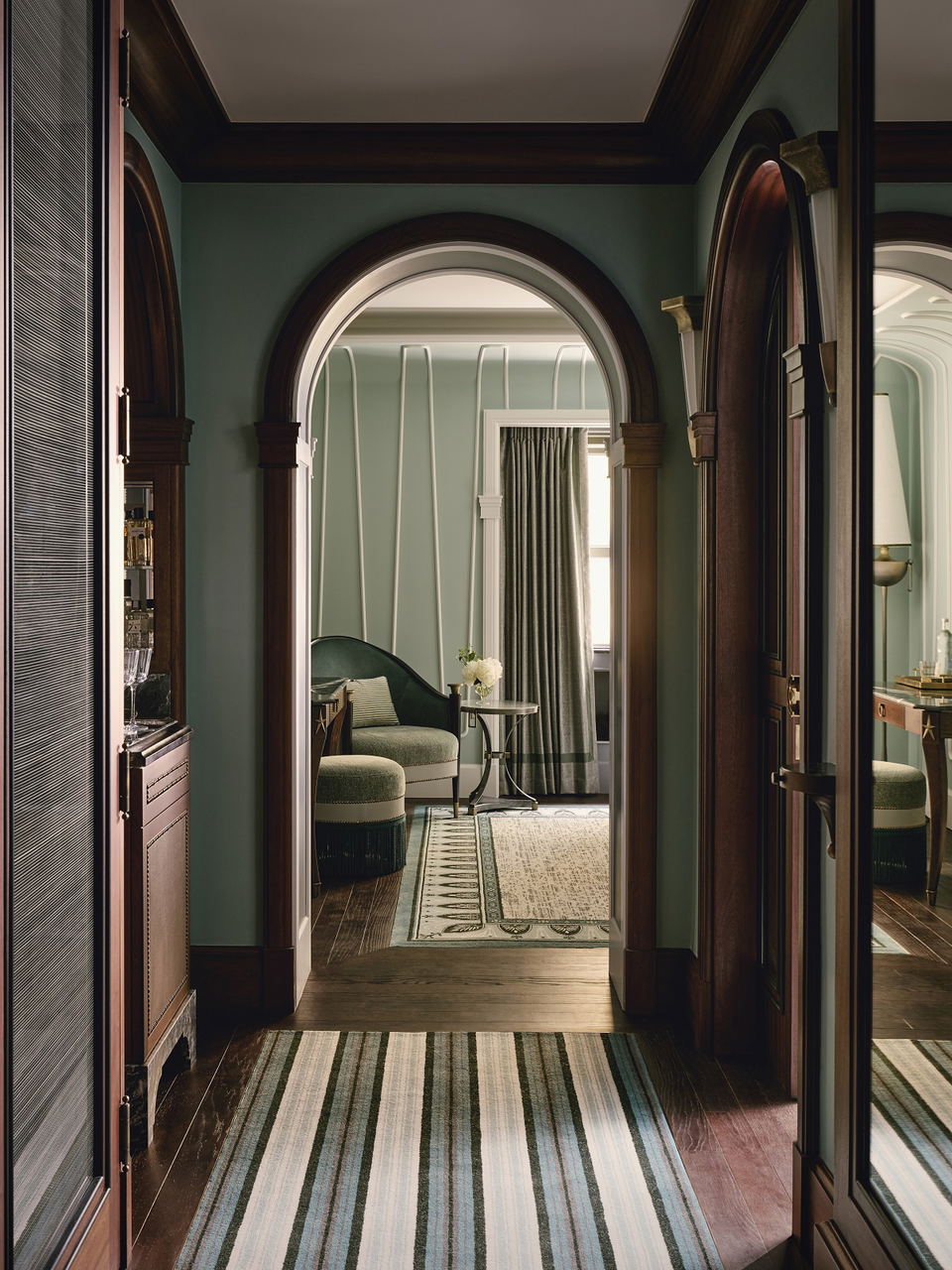 Interior hallway with mint green walls, dark wooden arched doorways, striped runner rugs, and a view into a bedroom with green furnishings and floor-to-ceiling curtains.