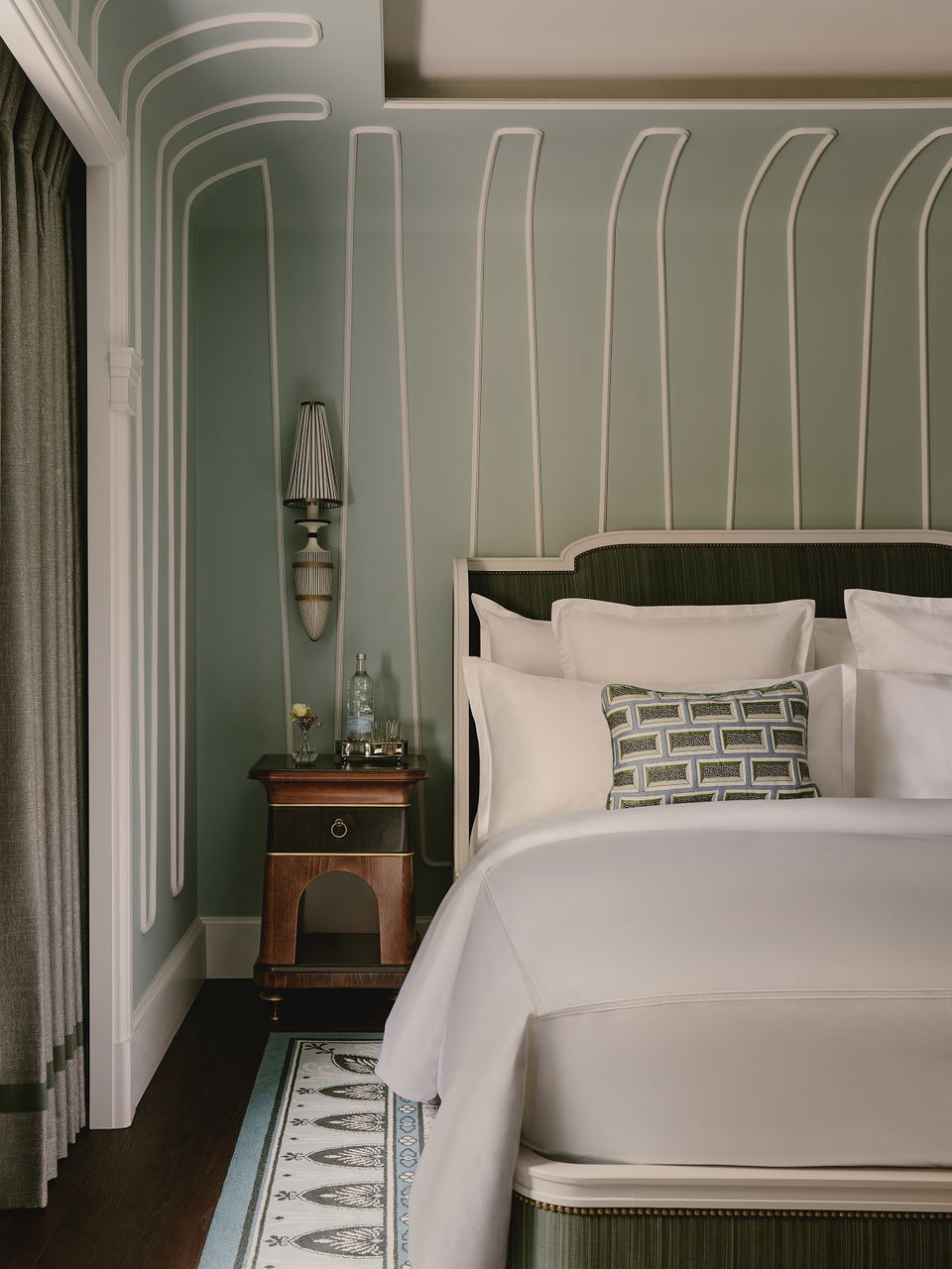 Modern bedroom with sage green walls, white vertical paneling, dark green bed headboard, white pillows, patterned throw pillow, wooden nightstand, and elegant wall sconce.