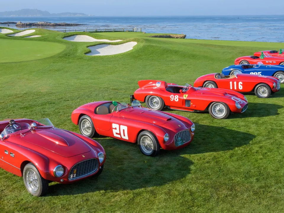 A line of classic red and blue Ferrari racing cars with numbers 20, 98, 116, and 225 on a green golf course next to the ocean.