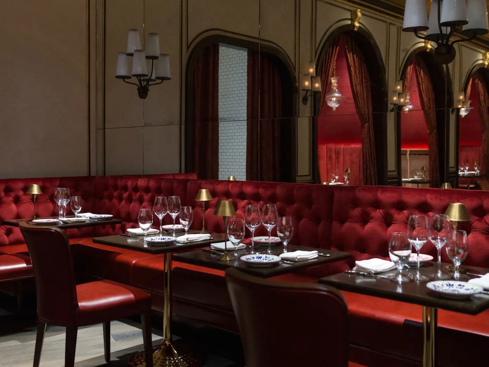 A luxurious dining room with deep red tufted velvet banquettes, dark wood tables set with wine glasses, and gold-accented arched mirrors.