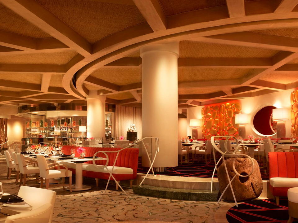 A warmly lit, upscale restaurant interior with red banquette seating, white chairs, and tables set with wine glasses and flowers. Ornate wooden beams