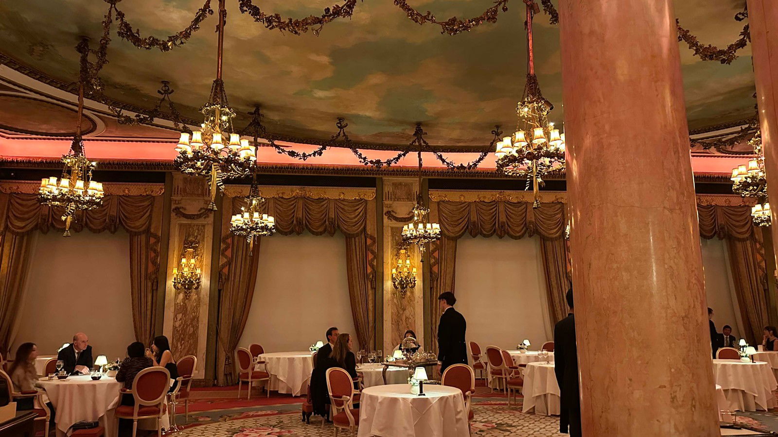 A Louis XVI-style dining room with ornate gold chandeliers, a painted sky ceiling, draped gold curtains, and white-tablecloth