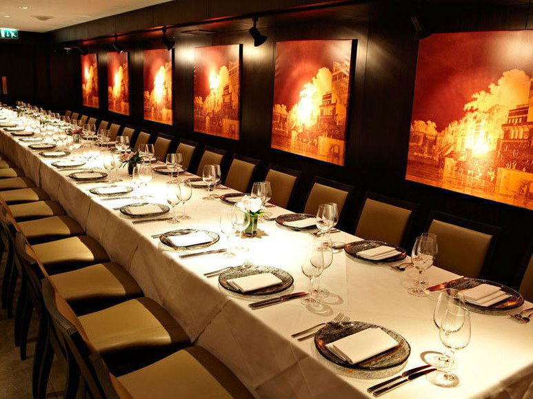 A long private dining room at Benares Mayfair with a white tablecloth, place settings, and amber-toned artwork on dark walls.