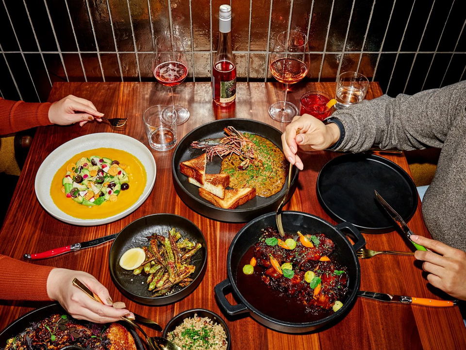 An overhead shot of two diners sharing multiple dishes at a restaurant table, including a shrimp dish, braised meat, roasted vegetables, and a salad,