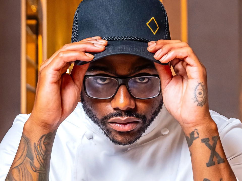 Chef, wearing a white chef's coat with his name embroidered, adjusts a black trucker cap while looking directly at the camera.