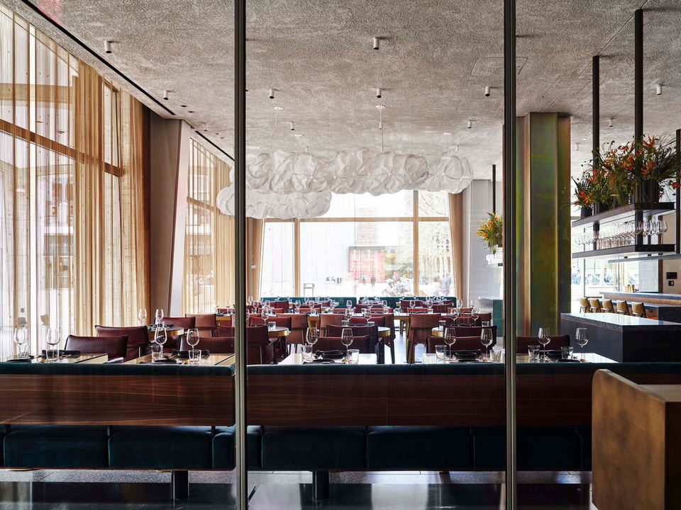A modern restaurant dining room with teal velvet banquettes, dark wood chairs, sheer golden curtains, and a cloud-like chandelier.