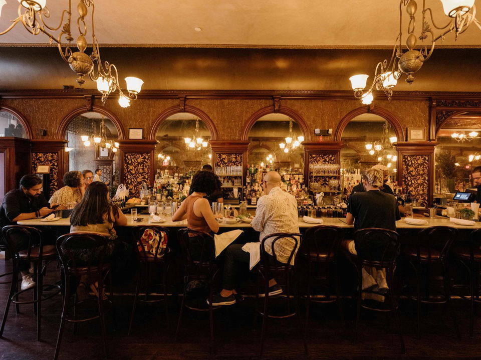 The interior of a bar with ornate Victorian-era decor, featuring arched mirrors, dark wood paneling, and brass chandeliers. Patrons sit on bentwood