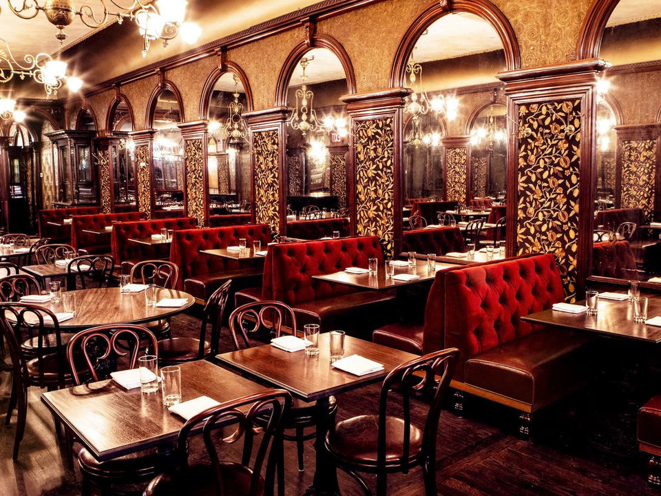 An ornate dining room with red velvet banquettes, dark wood tables, bentwood chairs, arched mirrors, and gold-leaf wallpaper.