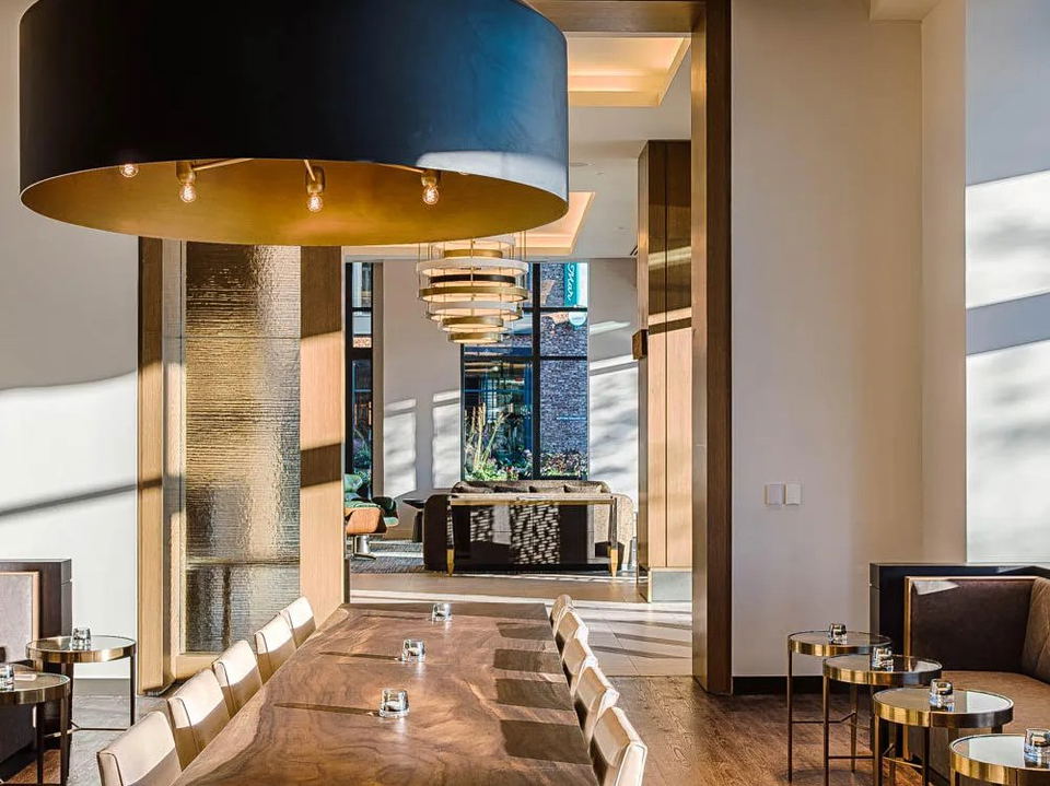 A modern dining area features a long wooden table, plush chairs, and striking black and gold light fixtures, bathed in natural light.